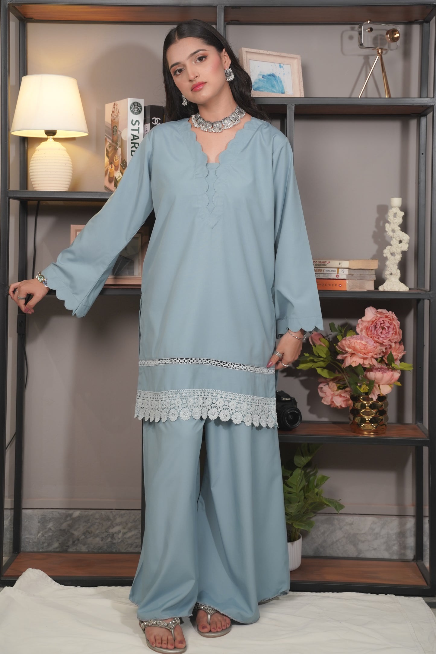 Woman in a light blue traditional outfit standing in front of a shelf with decorative items.