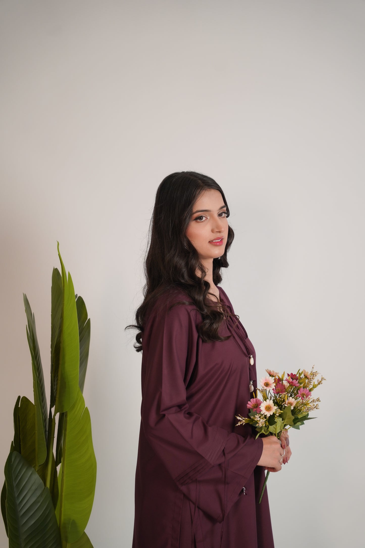 Woman in a purple dress holding flowers next to a plant against a plain background