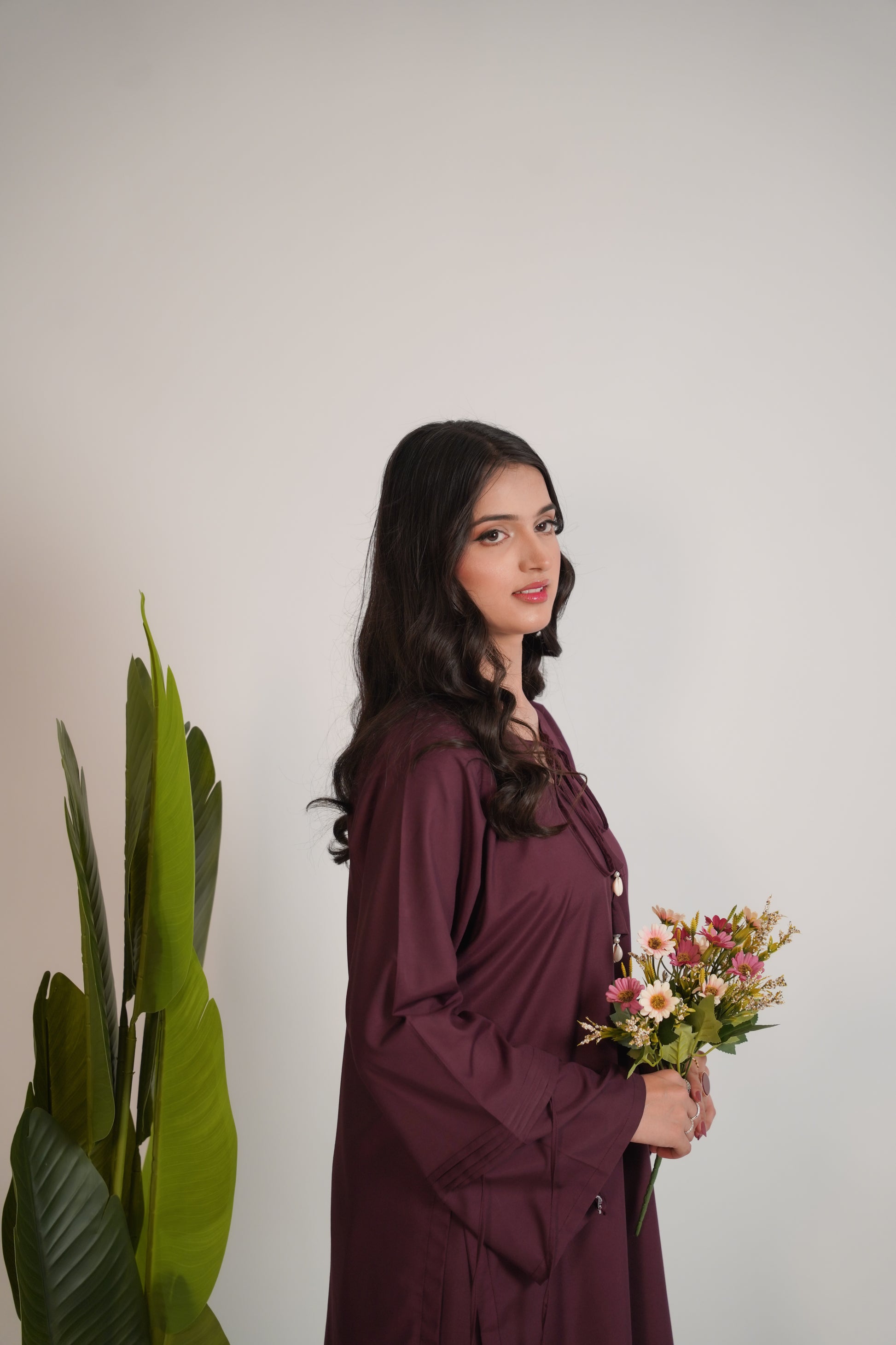 Woman in a purple dress holding flowers next to a plant against a plain background